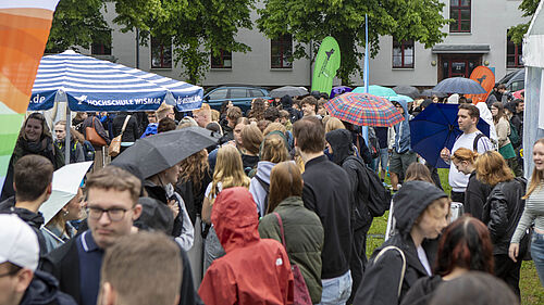 Es sind viele Menschen in Regenjacken und mit Schirmen zu sehen. Beachflags in Fakultätsfarben und ein großer Stand-Schirm mit der Aufschrift "Hochschule Wismar" ragen über die Köpfe der Personen hinaus.