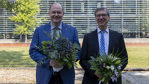 Die beiden Personen stehen nebeneinander auf dem Campus und halten je einen Blumenstrauß in den Händen.