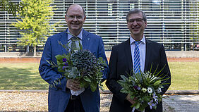 Die beiden Personen stehen nebeneinander auf dem Campus und halten je einen Blumenstrauß in den Händen.