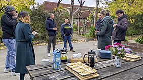 Gruppe von acht Personen steht im Schaugarten im Kreis, eine Person ist kaum zu sehen. Im Vordergrund steht ein Holztisch mit Kaffeekanne, Tellern, Kuchen und Milchpackung. Im Hintergrund sind herbstliche Bäume und Büsche zu sehen.