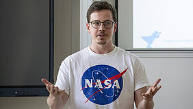 Ein junger Mann mit Brille und weißem T-Shirt steht vor der Projektionsfläche seiner Powerpoint-Präsentation. Mit offener Arm-Gestik steht er dem Publikum zugewandt.