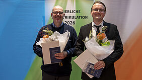 Zwei Männer in Anzügen stehen nebeneinander, halten jeweils einen Blumenstrauß und eine Urkunde, im Hintergrund ein Banner mit dem Text 'FEIERLICHE MATRIKULATION 2026'