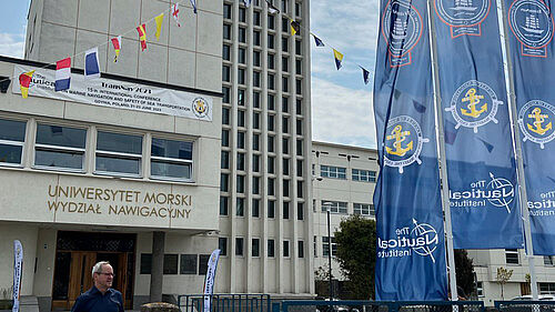 Es ist der Gebäudeeingang zu sehen. Rechts im Bild sind drei grroße Fahnen mit dem TransNav-Logo, links eine Beachflag und am oberen Rand etliche Signalflaggen.