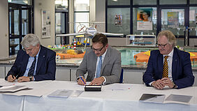 Die drei Herren sitzen an einem Tisch und unterzeichnen je ein Exemplar des VErtrages