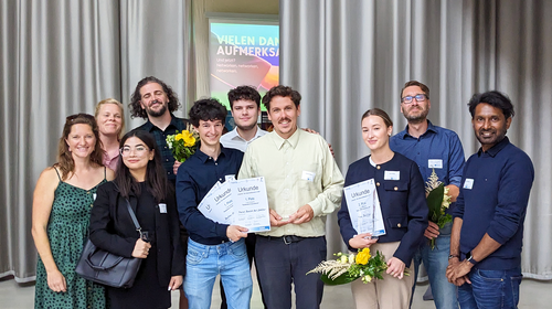Etwa zwölf Personen stehen nebeneinander in einem Raum. Vorne halten die Preisträger:innen der Hochschule Wismar Urkunden und Blumen. Dahinter stehen Mitglieder des Organisationsteams.