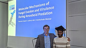 Der Professor steht neben dem Doktoranden und hat seinen linken Arm auf dessen Schulter gelegt. Nauman Saeed hat einen Doiktorhut auf. Hinter den beiden ist auf einer Präsentationsfolie der Titel der Arbeit zu sehen.