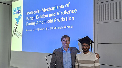 Der Professor steht neben dem Doktoranden und hat seinen linken Arm auf dessen Schulter gelegt. Nauman Saeed hat einen Doiktorhut auf. Hinter den beiden ist auf einer Präsentationsfolie der Titel der Arbeit zu sehen.