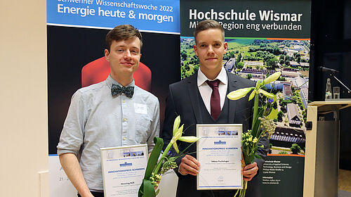 Zwei Preisträger stehen mit ihrem Blumenstrauß und der Urkunde in der Hand vor zwei Bannern, auf denen das Thema der Schweriner Wissenschaftswoche und der Campus der Hochschule Wismar zu sehen sind.