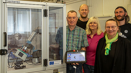 Die fünf Personen stehen rechts neben dem Gerät, das wie ein durchsichtiger Schrank aussieht, in dem sich ein Roboterarm befindet.