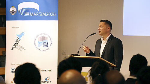 Mann im Anzug spricht an einem Rednerpult vor Publikum mit Banner im Hintergrund, auf dem 'MARSIM2026' und Logos zu sehen sind