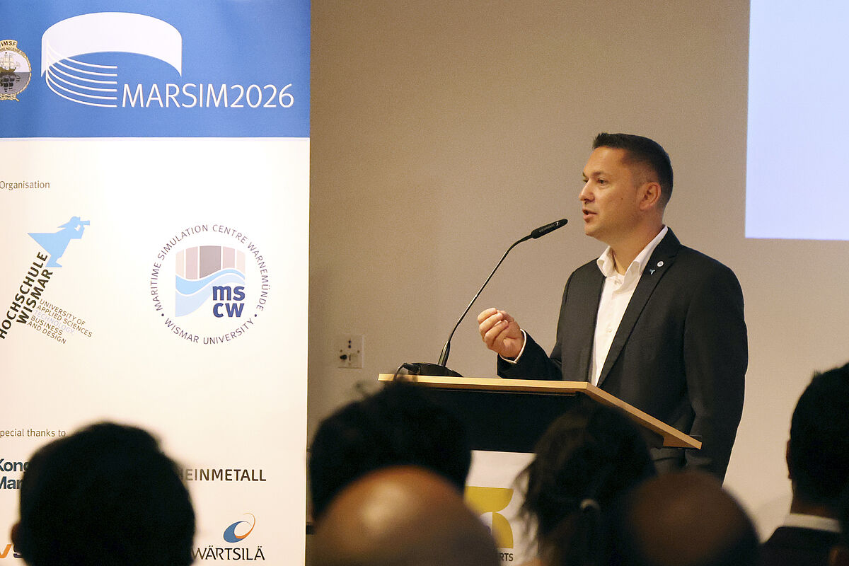 Mann im Anzug spricht an einem Rednerpult vor Publikum mit Banner im Hintergrund, auf dem 'MARSIM2026' und Logos zu sehen sind