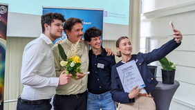 Vier junge Menschen, drei Männer und eine Frau, stehen zusammen und machen ein Selfie. Sie halten Blumen und Urkunden in den Händen und lachen in die Kamera. Es sind die Gewinnerteams der Hochschule Wismar.