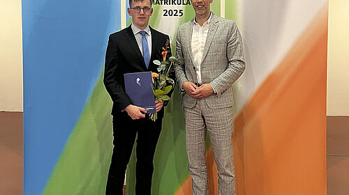Die beiden Persoenn stehen vor farbigen Bannern, auf denen "Feierliche Immatrikulation 2025" steht. Der Ausgezeichnete hält Blumen und Urkunde in der Hand.
