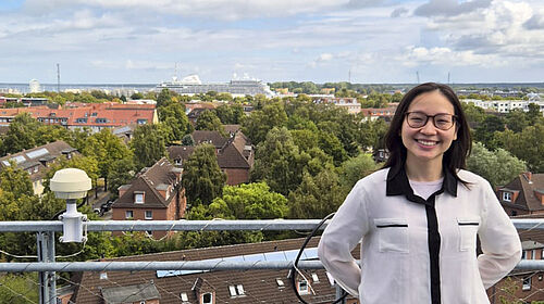 Eine Frau mit Brille und weißer Bluse steht lächelnd an einem Geländer mit Blick auf grüne Baumkronen, Wohnhäuser und einen Hafen mit Kreuzfahrtschiff im Hintergrund