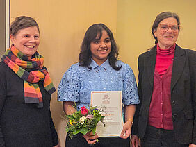 Die drei Frauen stehen nebeneinander und lächeln in die Kamera. Die Preisträgerin steht in der Mitte und hält eine Urkunde sowie einen Blumenstrauß in der Hand.