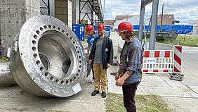 3 Studierende stehen vor dem Kernkraftwerk um ein großes Pumpenteil herum.