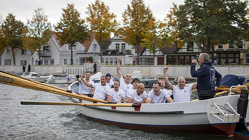 Männer in weißen T-Shirts sitzen im Kutter und reißen die Arme hoch.