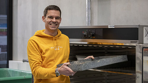 Der Professor steht vor einem Schrank, der wie ein großer Ofen aussieht. Er hält ein Blech in der Hand, das einem Backblech ähnlich sieht, und schiebt es in den Schrank.