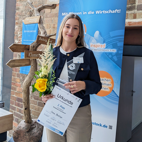 Eine junge Frau steht vor einem Wettbewerbsbanner und einem hölzernen Wegweiser. Sie hält eine Urkunde, einen Pokal und einen Blumenstrauß in der Hand und lächelt freundlich.