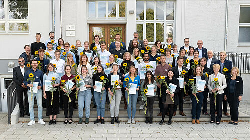 Es sind 53 Förderer sowie Deutschlandstipendiatinnen und -stipendiaten zu sehen, die gemeinsam auf der Seitentreppe des Hauses 1 stehen. Die Stipendiaten halten Sonnenblumenin den Händen.