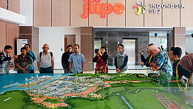 12 Personen stehen um ein großes Landschaftsmodell herum, das Grünflächen, Gebäude, einen Hafen und das Meer darstellt.