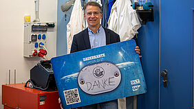 Der Mann steht in einem Labor und hält eine rund ein Meter breite Karte in der Hand. Sie sieht wie eine Dauerkarte aus, auf der in der Mitte "Danke" steht.