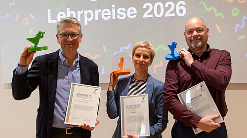 Die drei Personen stehen nebeneinander. Sie halten in der jeweils linken Hand eine Urkunde und  in Kopfhöhe einen kleinen Preispokal. Dieser hat jeweils die Farbe der Fakultät: Grün, Orange und Blau.