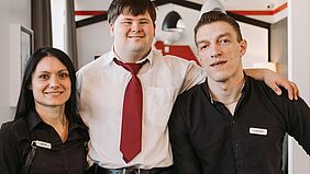 Drei junge Menschen lächeln in die Kamera. Zwei haben schwarze Hemden als Arbeitskleidung an und der junge Mann in der Mitte ein helles Hemd mit roter Krawatte. Er hat das Down-Syndrom.