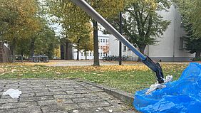 CampusCleanUp Mit einem Greifer wird ein herumliegendes Taschentuch in einen Müllbeutel gelegt. Im Hintergrund ist der herbstliche Campus zu erkennen.