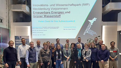 Die 21 Personen stehen in zwei REihen nebeneinander. Im Hintergrund ist das Präsentationsbild an der Wand zu erkennen.