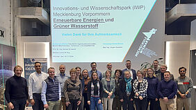 Die 21 Personen stehen in zwei REihen nebeneinander. Im Hintergrund ist das Präsentationsbild an der Wand zu erkennen.