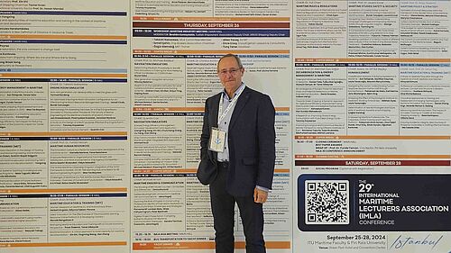 Die Person steht vor einer 2 Meter hohen Infowand, auf der das gesamte Tagungsprogramm abgebildet ist. Er trägt den Tagungs-Tag um den Hals.