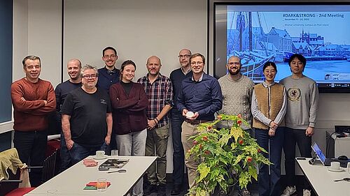 11 Personen stehen nebeneinander in einem Beratungsraum. Im Hintergrund ist eine Präsentation zu sehen.