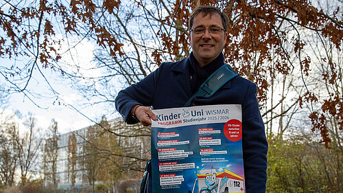 Professor Hillmann steht auf einer Wiese, die mit bunten Blättern zugedeckt ist. In den Händern hält er ein Plakat. Im Hintergrund sind Gebäude der Hochschule zu sehen.