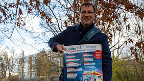Professor Hillmann steht auf einer Wiese, die mit bunten Blättern zugedeckt ist. In den Händern hält er ein Plakat. Im Hintergrund sind Gebäude der Hochschule zu sehen.
