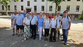 Eine Personengruppe steht bei sonnigem Wetter vor einem Gebäude.