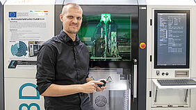Ein Mann in schwarzem Hemd steht vor einem Multimaterialdrucker, einem großem Gerät, einem Schrank ähnlich, mit beleuchtetem Innenraum und großem Bildschirm rechts.