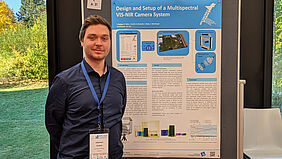 Ein Mann im schwarzen Hemd schaut in die Kamera, im Hintergrund ein blau gehaltenes, wissenschaftliches Plakat