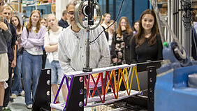 Man sieht eine bunte Brücke aus Papier, die in eine MEtallvorrichtung eingespannt ist. Dahinter sind unscharf Schülerinnen zu erkennen, die zuschauen.