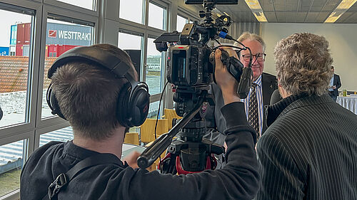 Zwei Männer sind von hinten zu sehen. Einer hat eine Kamera auf der Schulter, der andere ein Mikrofon in der Hand. Der Rektor schaut in die Kamera und spricht.