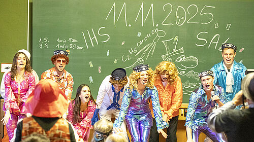 Studentinnnen und Studenten in Glitzerkostümen un mit Perücken tanzen auf der Bühne. Im Hintergrund an der Tafel steht mit Kreide "11.11.25, IHS, SAL"