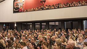 Publikum in einem Theater bei einem Festakt, sitzend und stehend auf zwei Ebenen. Sie blicken aufmerksam nach vornund manche klatschen.