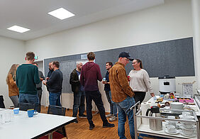 Teilnehmende stehen in kleinen Gruppen beieinander, sprechen miteinander und halten Becher mit Punsch. Auf den Tischen stehen Teller mit Gebäck.