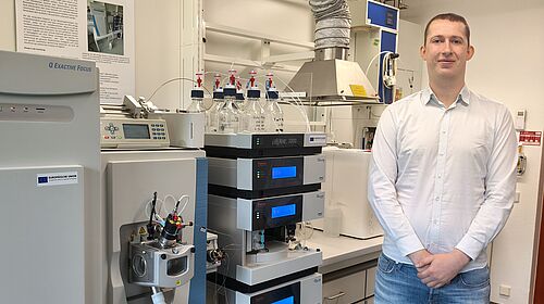 Der junge Mann trägt ein weißes Hemd und eine Jeans. Er steht in einem Labor neben Apparaturen. Im Hintergrund sind weitere Labormöbel und technische Geräte zu sehen.