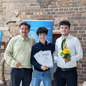 Drei junge Männer stehen vor einer Backsteinwand. Sie halten Urkunden, einen Pokal und einen Blumenstrauß in den Händen und lächeln stolz in die Kamera.