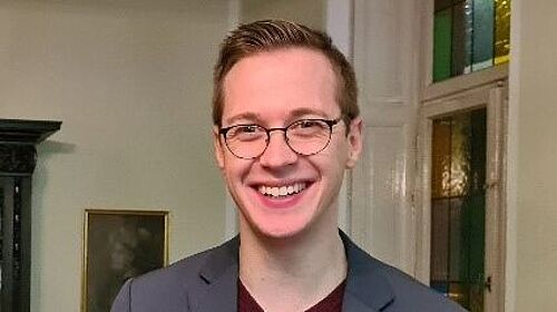 Junger Mann im Anzug und mit Brille lacht freundlich, im Hintergrund ein Raum mit bunten Fenstern, Bild und schönem alten Schrank..