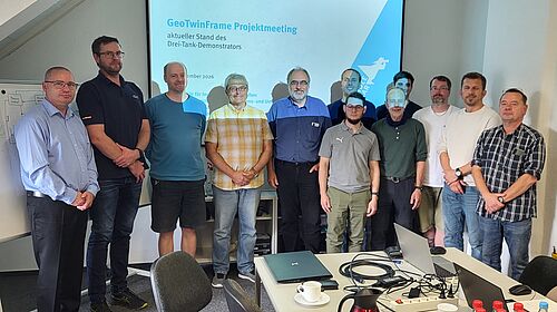 12 Männer stehen in einem Seminarraum vor einem Screen mit dem Titel "GeoTwinFrame Projektmeeting"