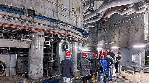 Mehrere Personen mit Schutzhelmen laufen entlang einer großtechnischen Industrieanlage.
