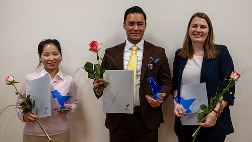 Die drei Personen stehen nebeneinander. Sie halten ihre Urkunde, den Preis und eine Rose in der Hand.