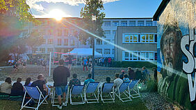 Im Vordergrund ist das Publikum auf dem Rasen und in Liegestühlen sitzend von hinten zu sehen. Sie verfolgen ein Spiel auf dem Volleyballfeld vor ihnen. 